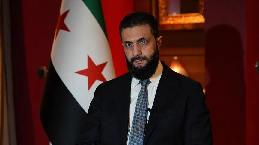 Syrian President Ahmad al-Sharaa, bearded and in a dark suit with a lapel microphone, seated for an interview in front of the Syrian flag.