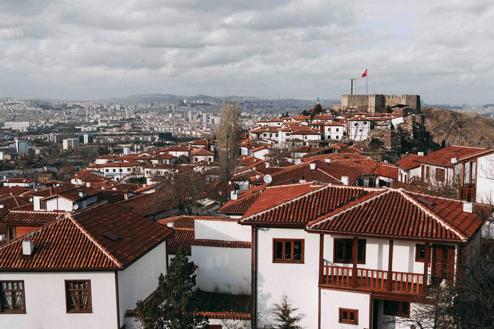View across the historic Hisar quarter of Ankara — the white-walled, red-tiled traditional houses climbing toward the Ankara Kalesi (citadel) flying the Turkish flag, with the modern city extending beyond under a cloudy plateau sky