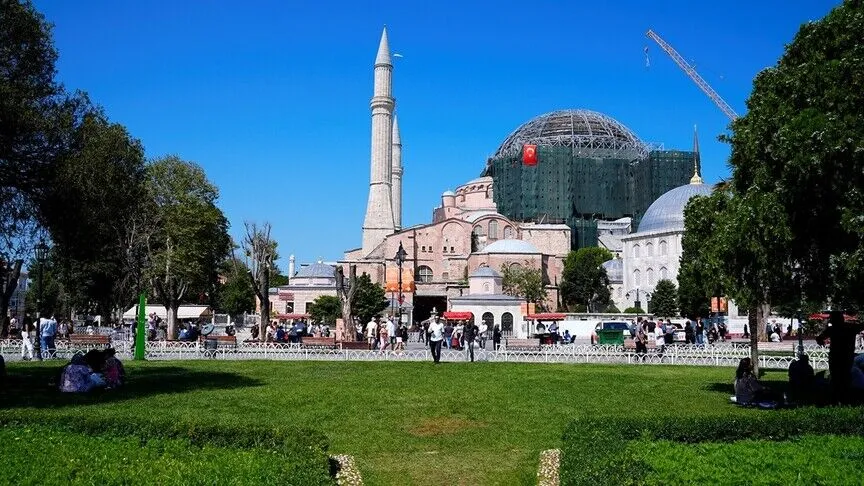Hagia Sophia seen across the green of Sultanahmet, its main dome encased in restoration scaffolding and mesh with a construction crane behind, visitors on the lawn in the foreground.