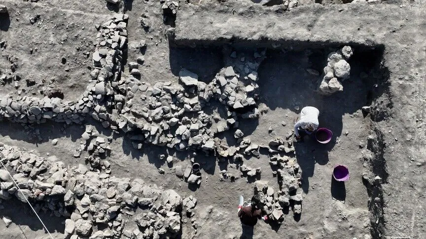 An archaeological excavation photographed from directly above, showing exposed stone wall foundations, cut trenches and two workers with buckets and hand tools.