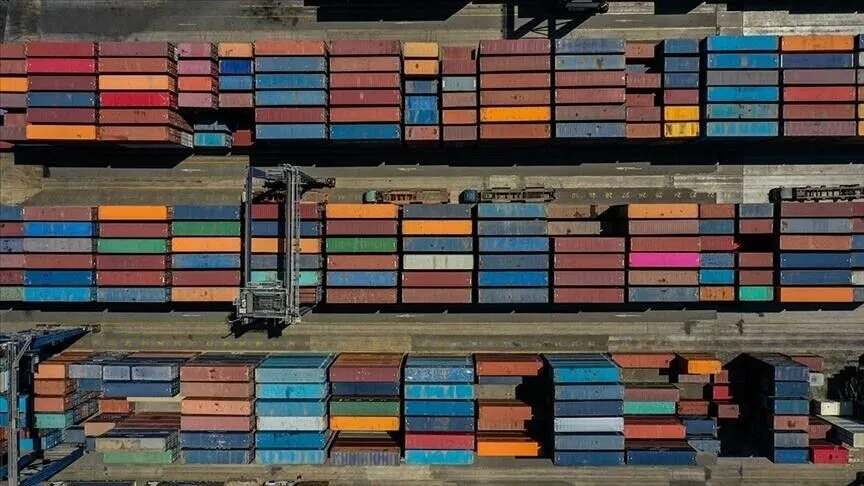 Aerial view of thousands of stacked shipping containers in rows at a port, with a straddle carrier moving between them.