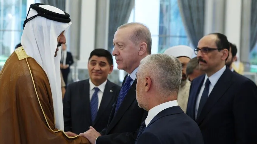 President Erdoğan clasps hands with a Qatari official in traditional dress in a reception hall, members of his delegation standing behind him.