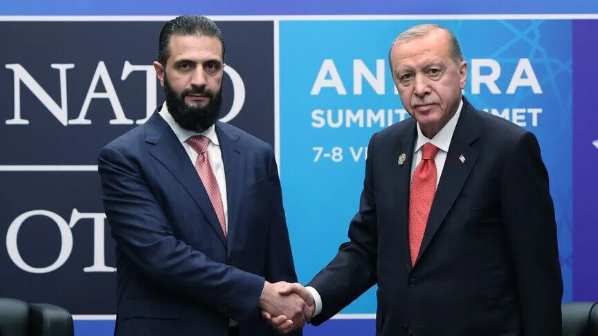 Ahmad al-Sharaa and Recep Tayyip Erdoğan shake hands in front of a backdrop reading NATO Ankara Summit, both in dark suits.