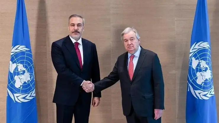 Hakan Fidan and António Guterres shake hands in front of two United Nations flags, both in dark suits.