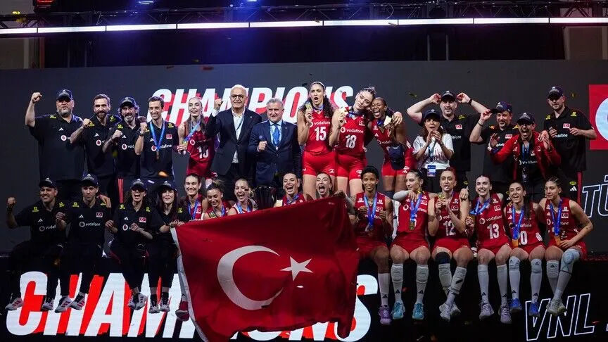 Türkiye's women's volleyball team and staff on the podium behind a large Turkish flag, wearing medals under a screen reading CHAMPIONS, with confetti in the air.