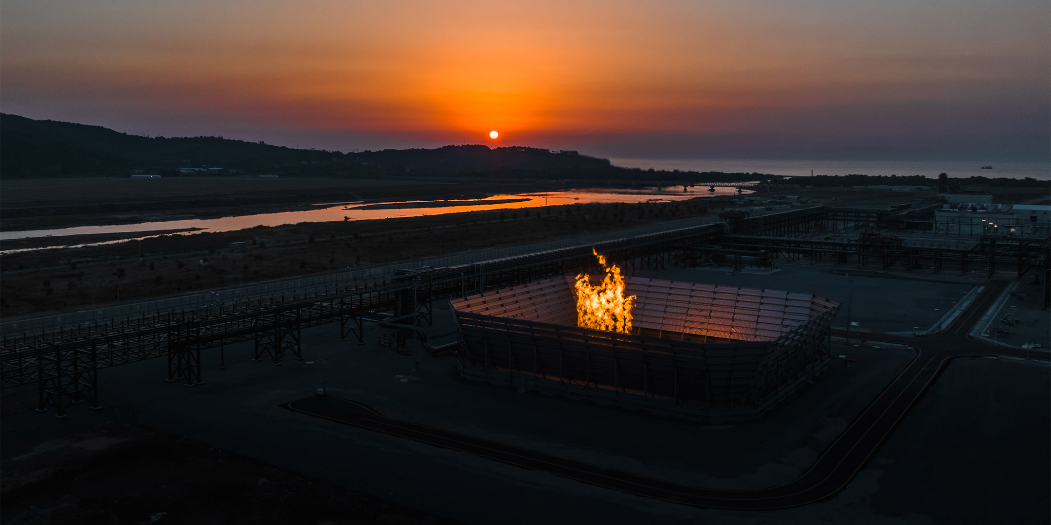 Aerial view of the Filyos Natural Gas Processing Terminal at sunset, with the project's gas flare burning beside the processing trains; the Filyos River and the Black Sea coast in the distance.