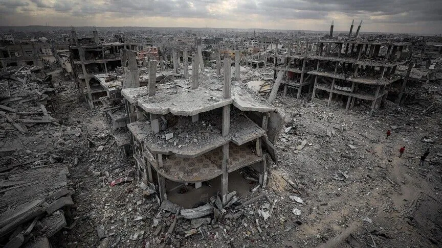 An aerial view of a destroyed district of Gaza, where a collapsed multistory building stands in the foreground amid rubble stretching to the horizon, with a few people walking through it.
