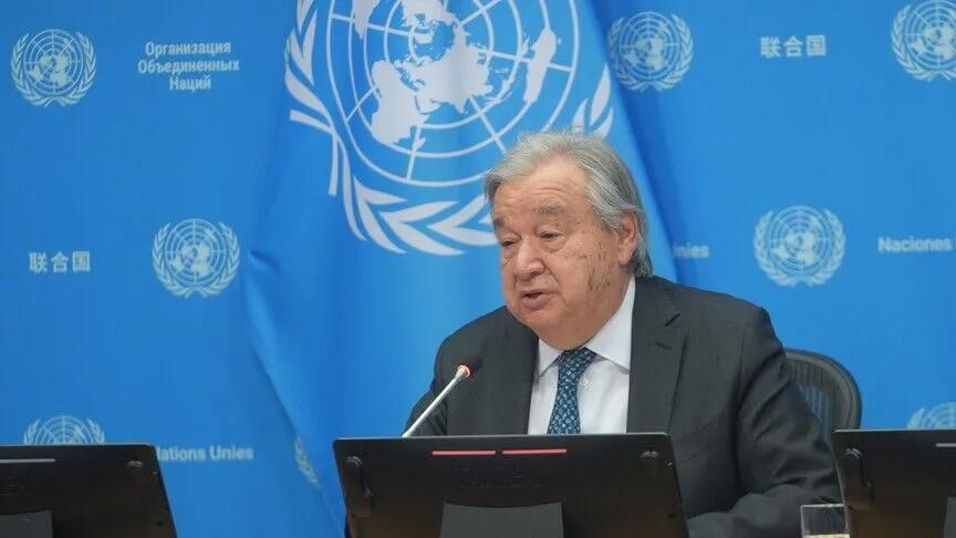 UN Secretary-General António Guterres speaks into a microphone in front of the United Nations flag, with a backdrop showing the organization's name in several languages.