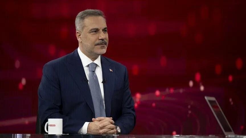 Foreign Minister Hakan Fidan seated in a television studio during an interview, a TRT Haber mug on the desk.