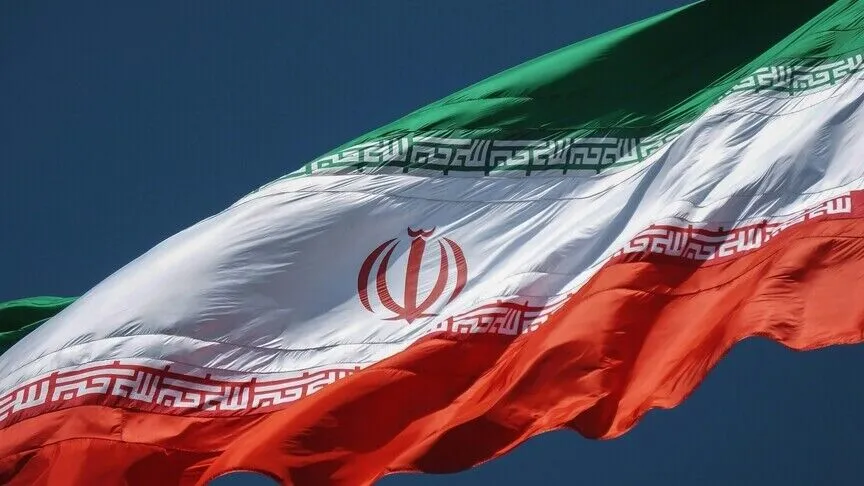 The flag of Iran, green, white and red with the national emblem at its center, filling the frame against a clear blue sky.