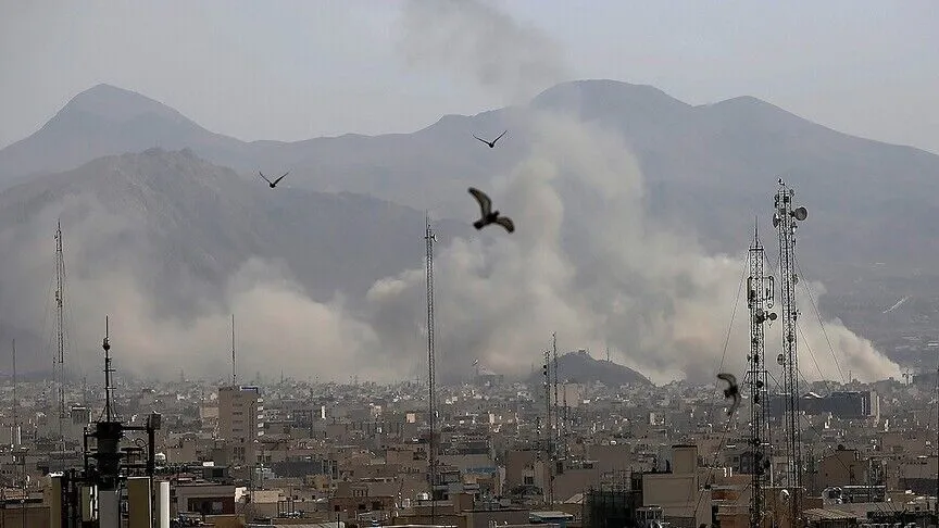 Smoke rises over a city skyline in Iran, with birds in flight and telecom towers in the foreground and mountains behind.