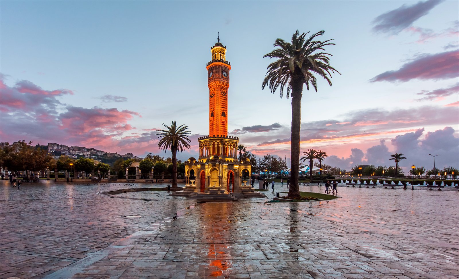 The İzmir clock tower (Saat Kulesi) in Konak Meydanı at dusk, palm trees against a pink-and-blue sky, the wet square reflecting the lit Ottoman-revival ironwork