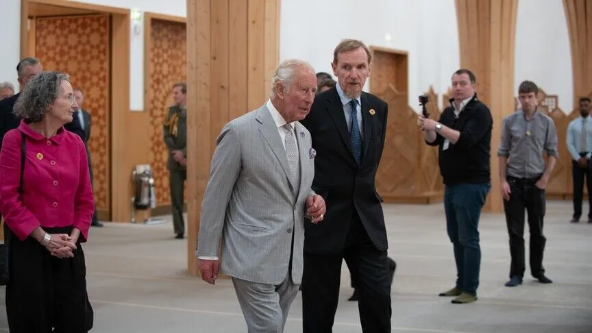King Charles III, in a grey suit, walks through the timber-columned interior of Cambridge Central Mosque with a guide, past its geometric-patterned wooden screens.