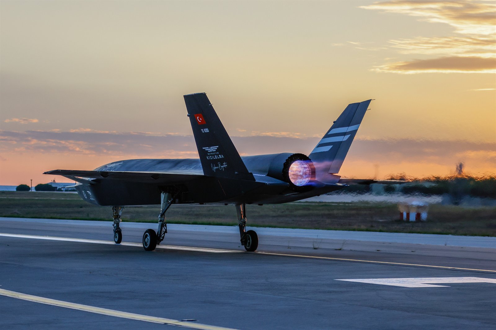 The Bayraktar Kızılelma unmanned fighter jet on a runway at dusk, afterburner lit, its twin tail carrying the Turkish flag.