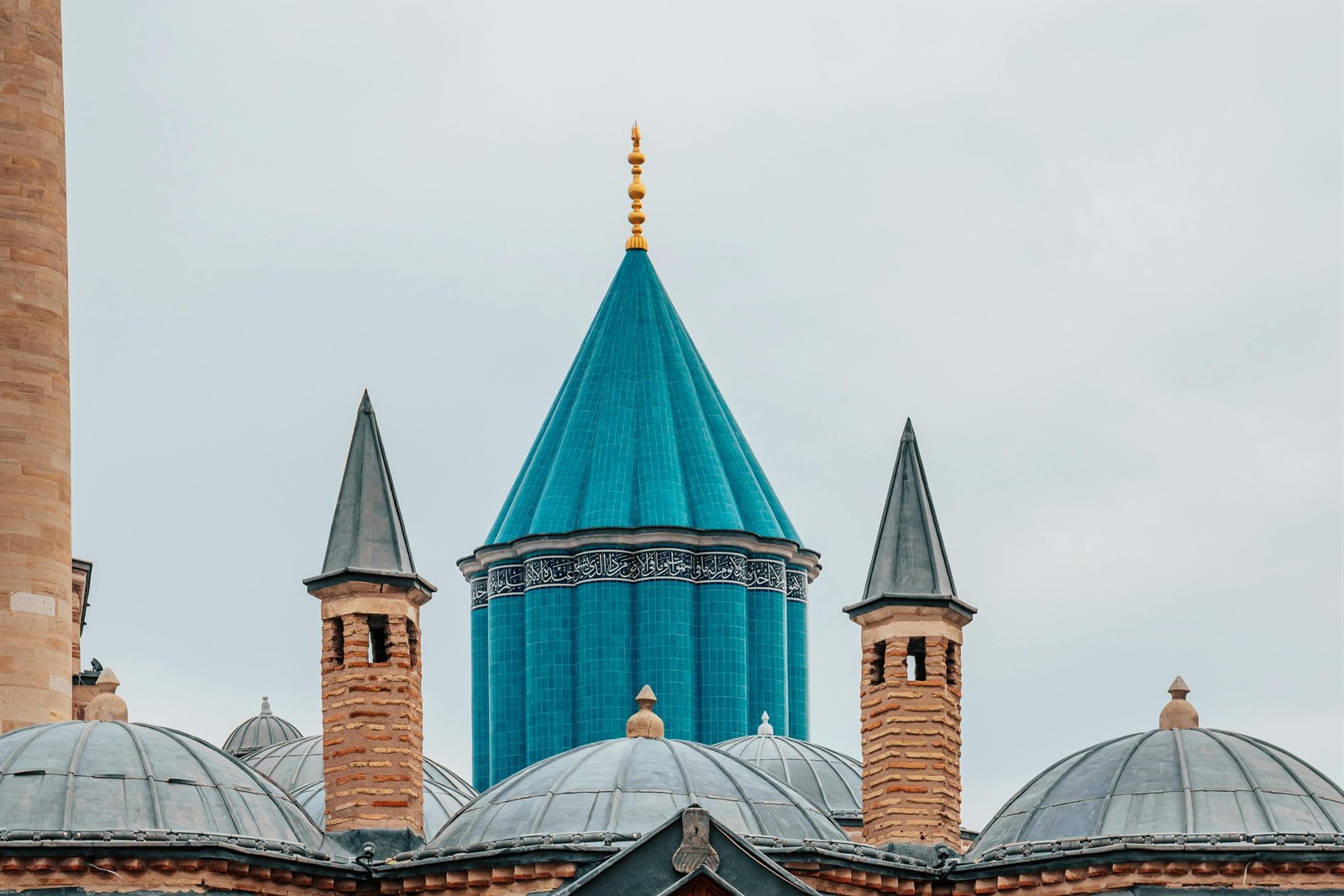 The Mevlâna Müzesi in Konya — the turquoise-tiled conical dome (yeşil türbe) of the Mevlâna Celâleddin-i Rûmî mausoleum rising above the lead-domed dergâh complex and its stone-walled courtyard