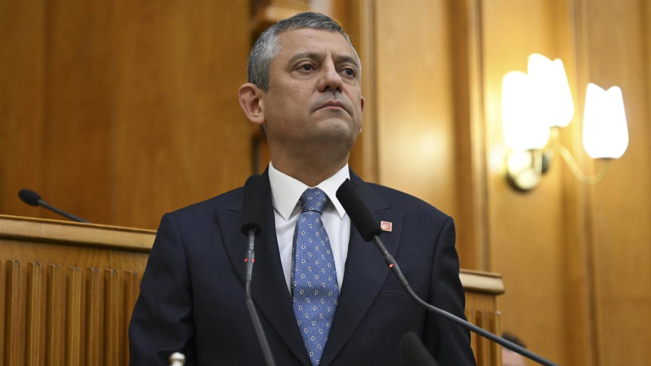 Özgür Özel, in a dark suit with a CHP lapel pin, stands at a microphone-lined lectern in parliament, looking ahead with a serious expression.