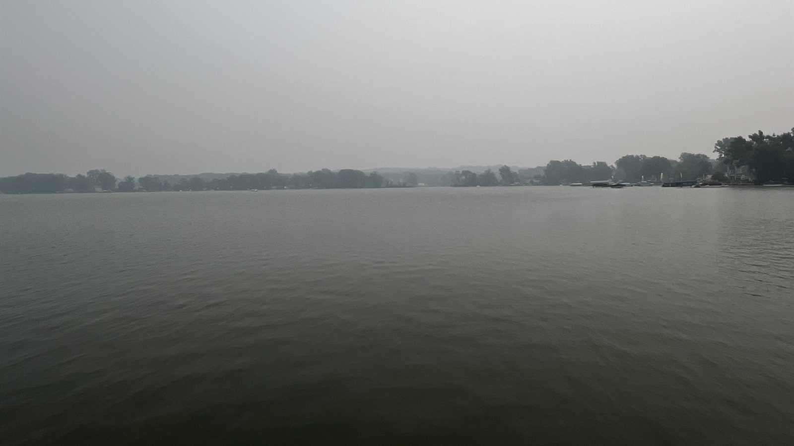 Grey wildfire haze hanging low over a lake and treeline in the Great Lakes region.