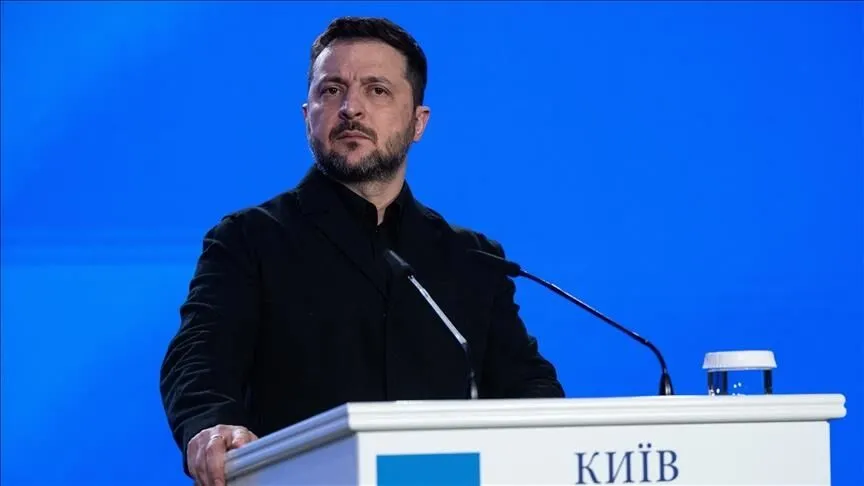 Volodymyr Zelenskyy in a black jacket stands at a lectern marked Kyiv in Ukrainian, speaking into microphones against a blue backdrop.