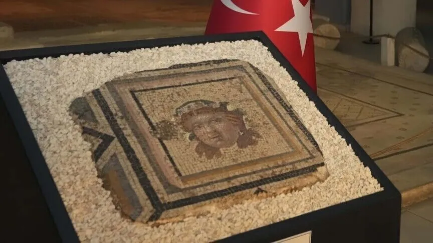 A framed panel of the Zeugma mosaic, a wreathed face worked in fine stone tesserae, displayed on a plinth in white gravel with a Turkish flag behind it.